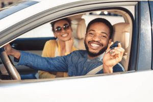 Visiting car dealership. Afro couple showing car key, looking at camera and smiling, sitting in new car